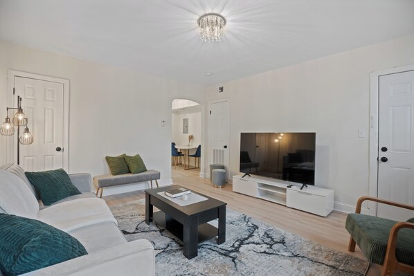 A modern living room with a sofa, coffee table, and TV, featuring a chandelier, white walls, and a wooden floor. - Located in the heart of SE Washington, DC. Perfect for both short /long stays. (Washington)