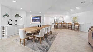A spacious dining area with a long wooden table and chairs, a kitchen with white cabinets, and a large window.