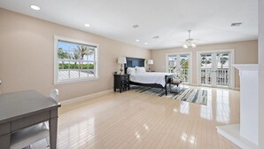 A spacious bedroom with a large bed, a nightstand, a desk, and a balcony with glass doors.