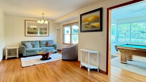 A spacious living room with hardwood flooring, a grey sofa, a coffee table, a white radiator, and a pool table. - Poseidon’s Big Getaway – 3BR/2BA Surrounded by Nature, Minutes from the Beach! (Kitty Hawk)