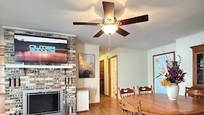 A spacious dining area with a wooden table and chairs, a fireplace with a TV above it, and a ceiling fan. - Poseidon’s Big Getaway – 3BR/2BA Surrounded by Nature, Minutes from the Beach! (Kitty Hawk)