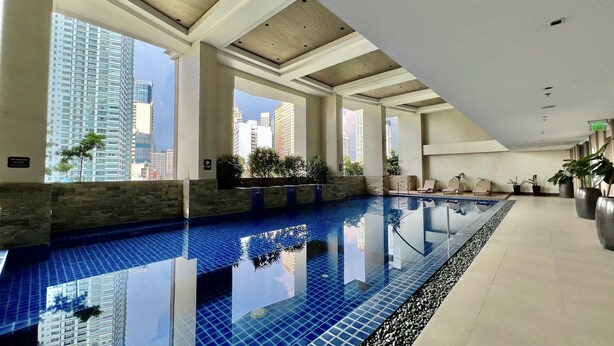 A modern indoor swimming pool with clear views of tall buildings, large windows, and a calm water surface. - KL Tower - Cozy 1BR Deluxe with Balcony near Greenbelt Mall (Makati)