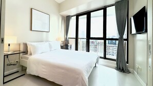 A modern hotel room with a large bed, a flat-screen TV, a city view, and a window with curtains. - KL Tower - Cozy 1BR Deluxe with Balcony near Greenbelt Mall (Makati)