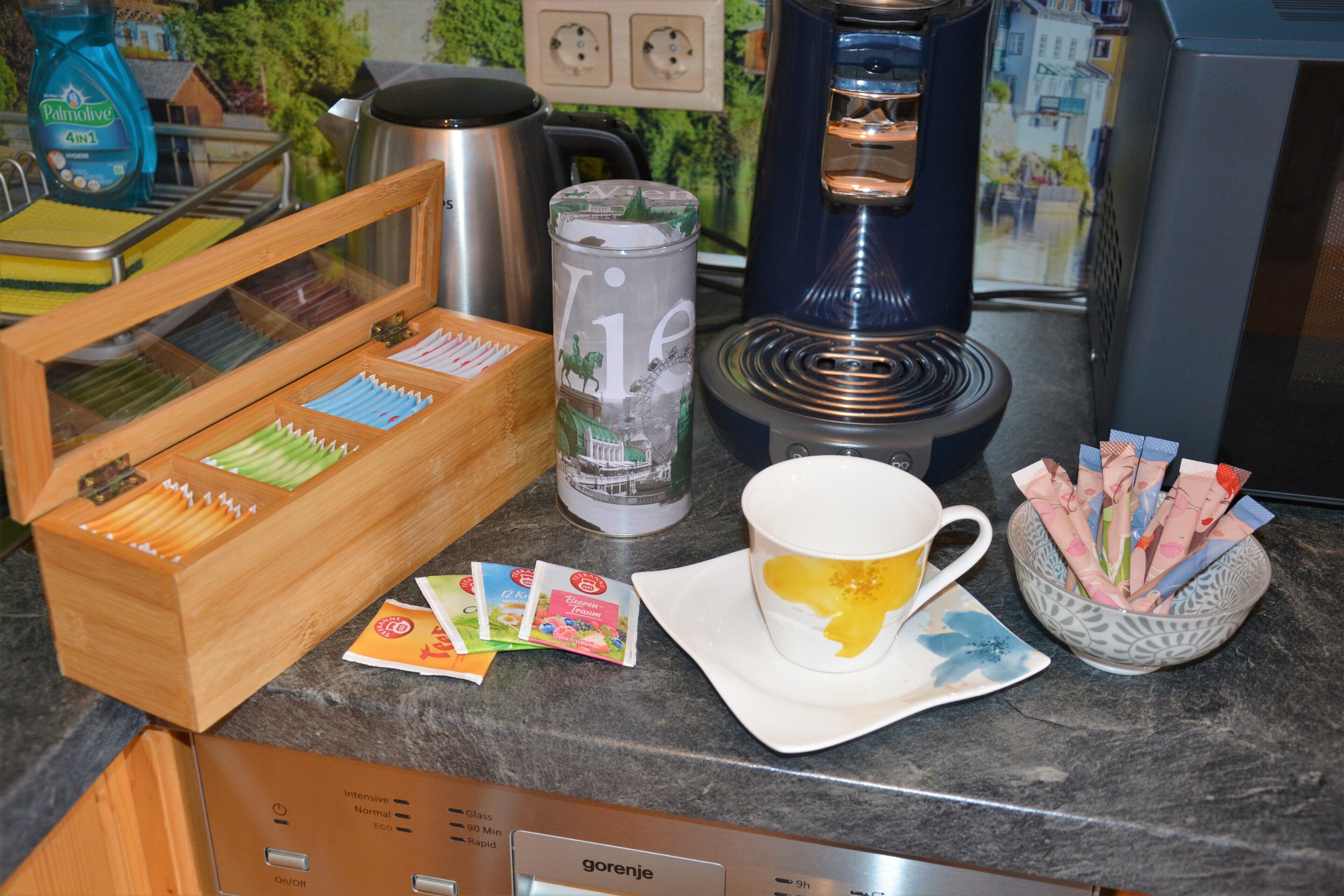 A coffee machine, a cup of coffee, a thermos, and a wooden box with tea bags on a kitchen counter.