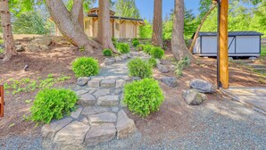 A wooden cabin with a stone pathway leading to a wooden deck, surrounded by trees and a small shed.