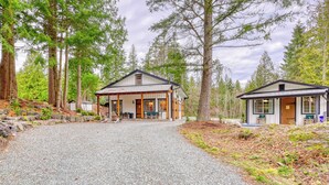 A two-story lodge with a covered patio and a separate cabin, surrounded by tall trees and a gravel driveway.