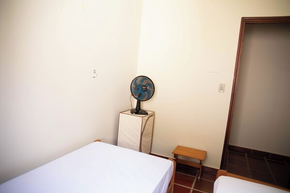 A small, simple room with a single bed, a small table, and a standing fan. - Casa Águas da Prata (Aguas da Prata)