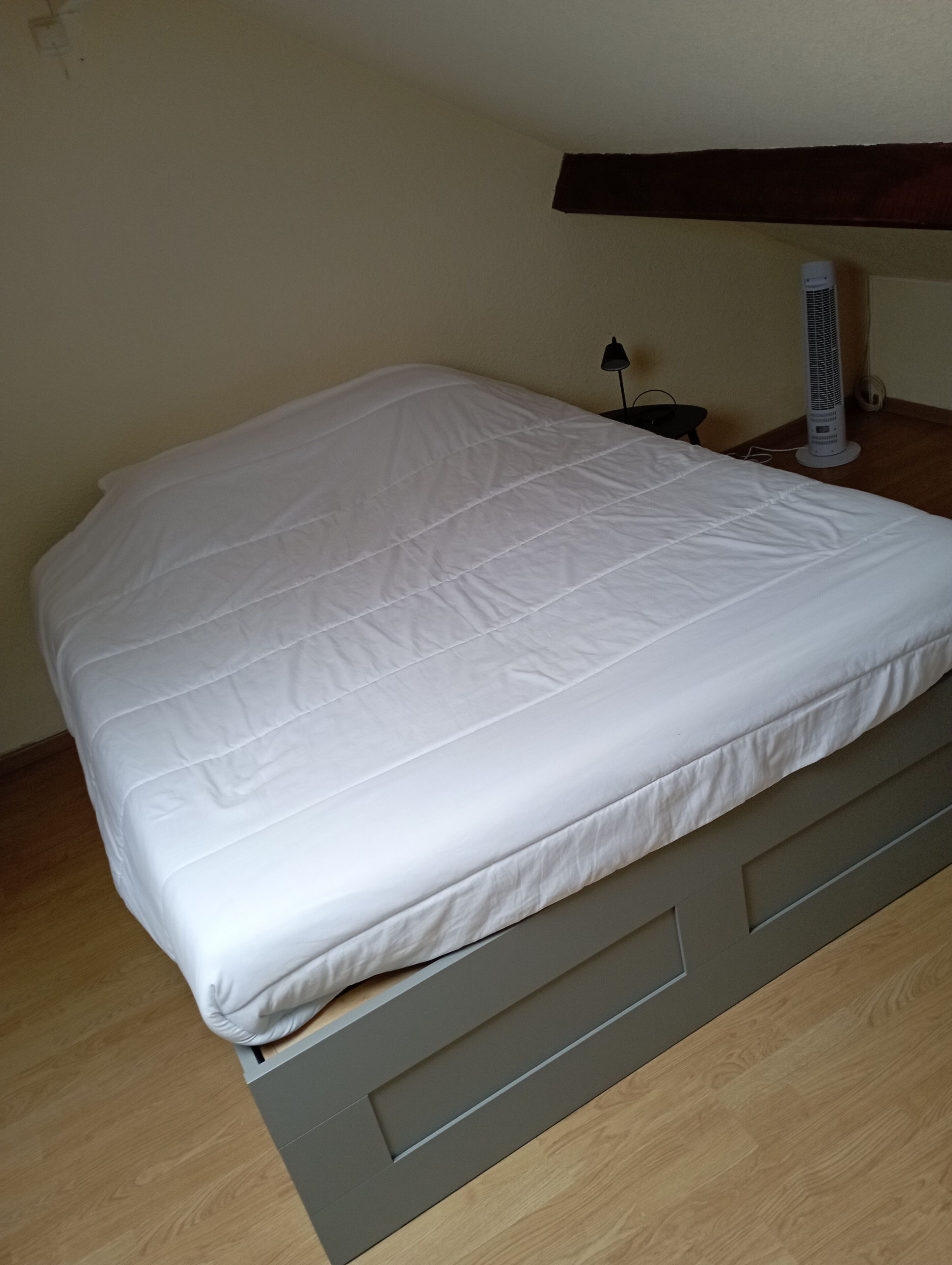 Un lit avec un matelas blanc et une tête de lit grise, dans une pièce dotée d’un plancher en bois et d’un climatiseur fixé au mur.