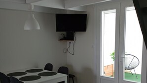 En matplats med ett vitt bord och svarta stolar, en väggmonterad TV och en dörr som leder ut till en balkong med en stol.