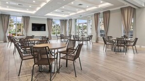 A spacious dining area with multiple tables and chairs, large windows with curtains, and a flat-screen TV mounted on the wall. - 2-bedroom condo, 5 min to private beach, golf, tiki new pool, hot tub fitness rm (Fort Pierce)