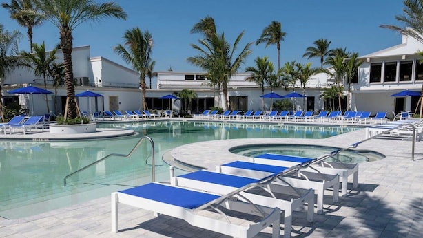 A pool area with lounge chairs and umbrellas, surrounded by palm trees and a building with large windows. - 2-bedroom condo, 5 min to private beach, golf, tiki new pool, hot tub fitness rm (Fort Pierce)
