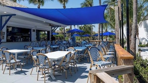 An outdoor seating area with tables and chairs, a bar, and palm trees in the background. - 2-bedroom condo, 5 min to private beach, golf, tiki new pool, hot tub fitness rm (Fort Pierce)