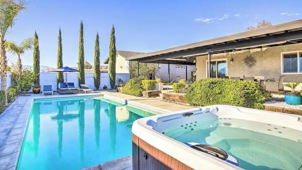 A swimming pool with lounge chairs and a hot tub, surrounded by tall trees and a building with a covered patio.