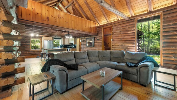 A log cabin interior with a large gray sectional sofa, a wooden coffee table, and a dining area with a kitchen in the background.