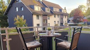 A balcony with two wicker chairs and a small round table, overlooking a residential area with houses and trees. - Harzblick apartment - your retreat in the middle of nature (Braunlage)