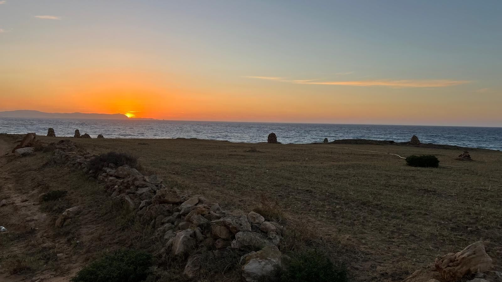 Un paisaje costero con atardecer, terreno rocoso y vegetación escasa.