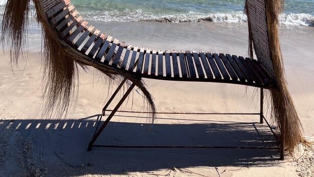 A beach chair with a thatched roof, positioned on a sandy beach with the ocean in the background.