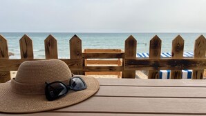 A wooden table with a straw hat and sunglasses on it, overlooking a beach with a wooden fence and striped beach chairs in the background.