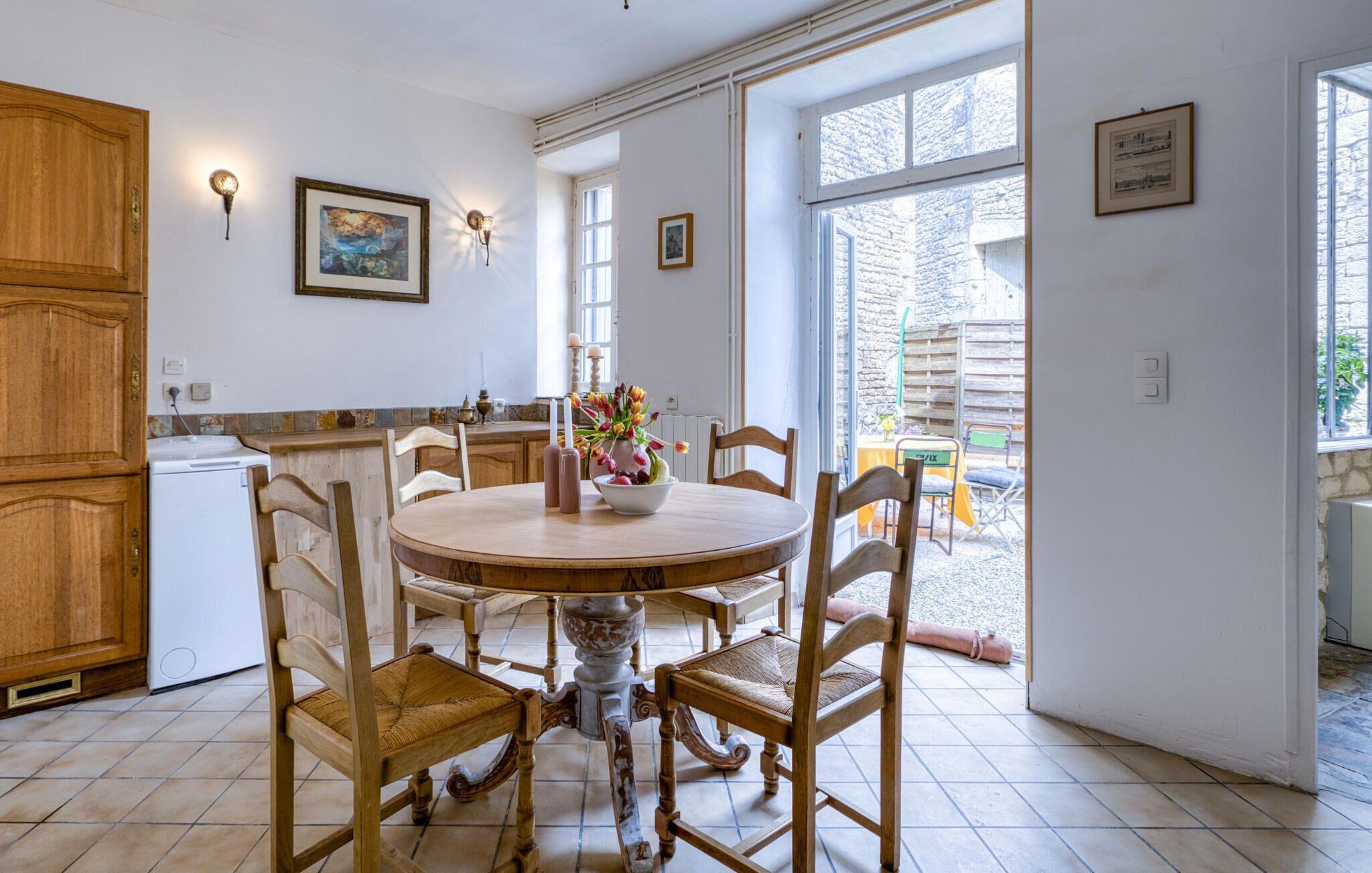 A kitchen with a round wooden table, chairs, and a white refrigerator. There are two wall-mounted light fixtures and a window with a view of the outside.
