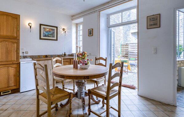 A kitchen with a round wooden table, chairs, and a white refrigerator. There are two wall-mounted light fixtures and a window with a view of the outside.