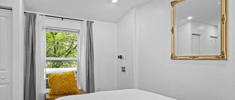 A bedroom with a bed, a window with curtains, a mirror, and a door.