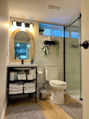 A modern bathroom with a glass shower enclosure, a white toilet, a sink with a mirror, and a shelf with towels and toiletries.