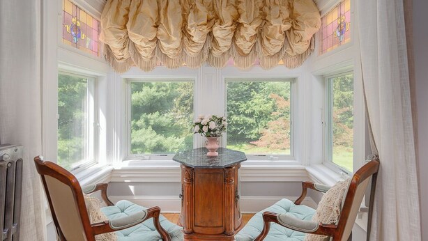 A room with a bay window, a wooden table with a vase of flowers, and two upholstered chairs.