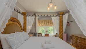 A large bed with white linens, a wooden headboard, and a canopy. There is a small table with a vase of flowers and a chair in the corner.