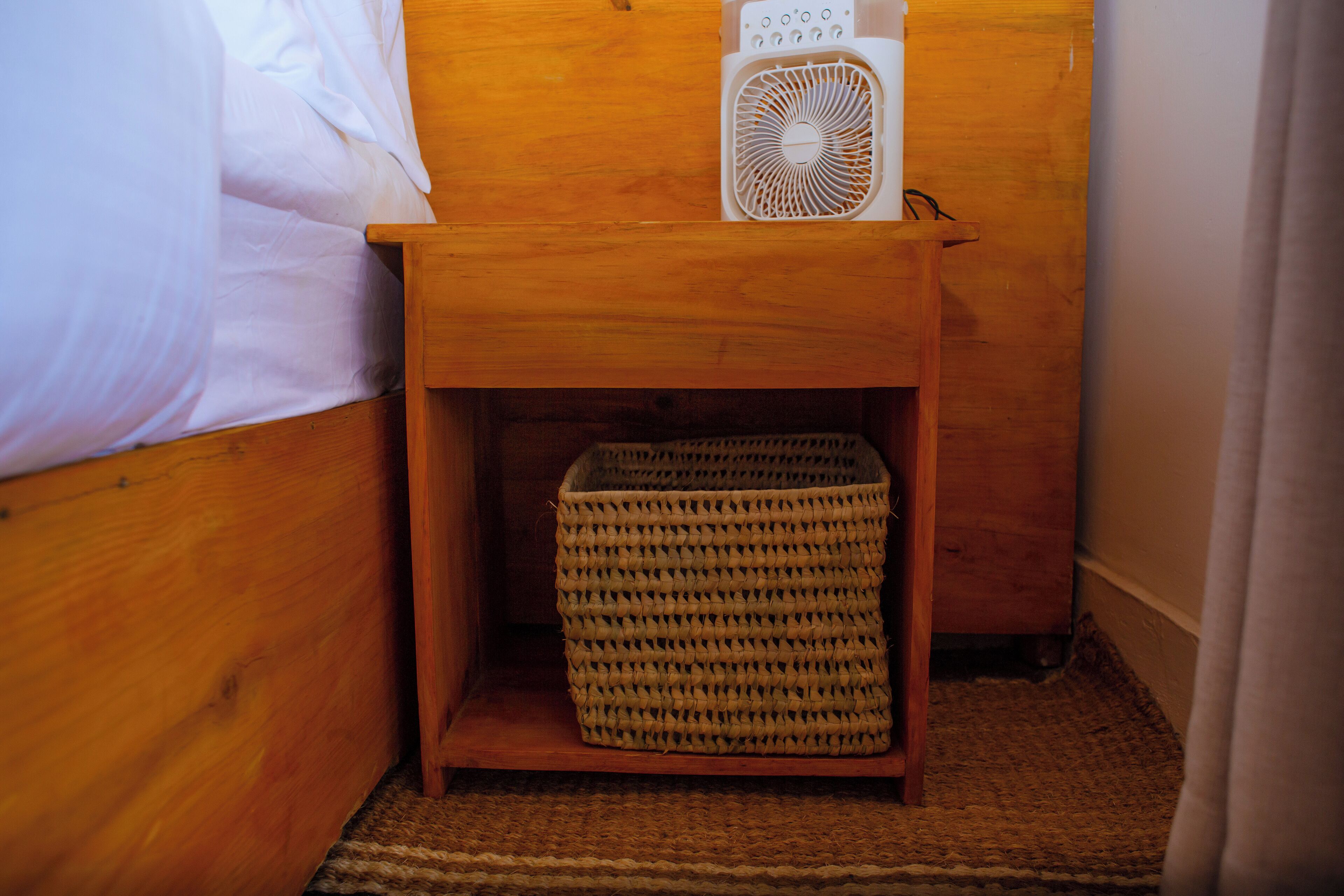 Una mesita de noche de madera con una cesta tejida debajo, un ventilador blanco encima y una cama con ropa de cama blanca.