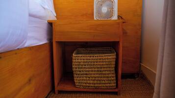 Una mesita de noche de madera con una cesta tejida debajo, un ventilador blanco encima y una cama con ropa de cama blanca.