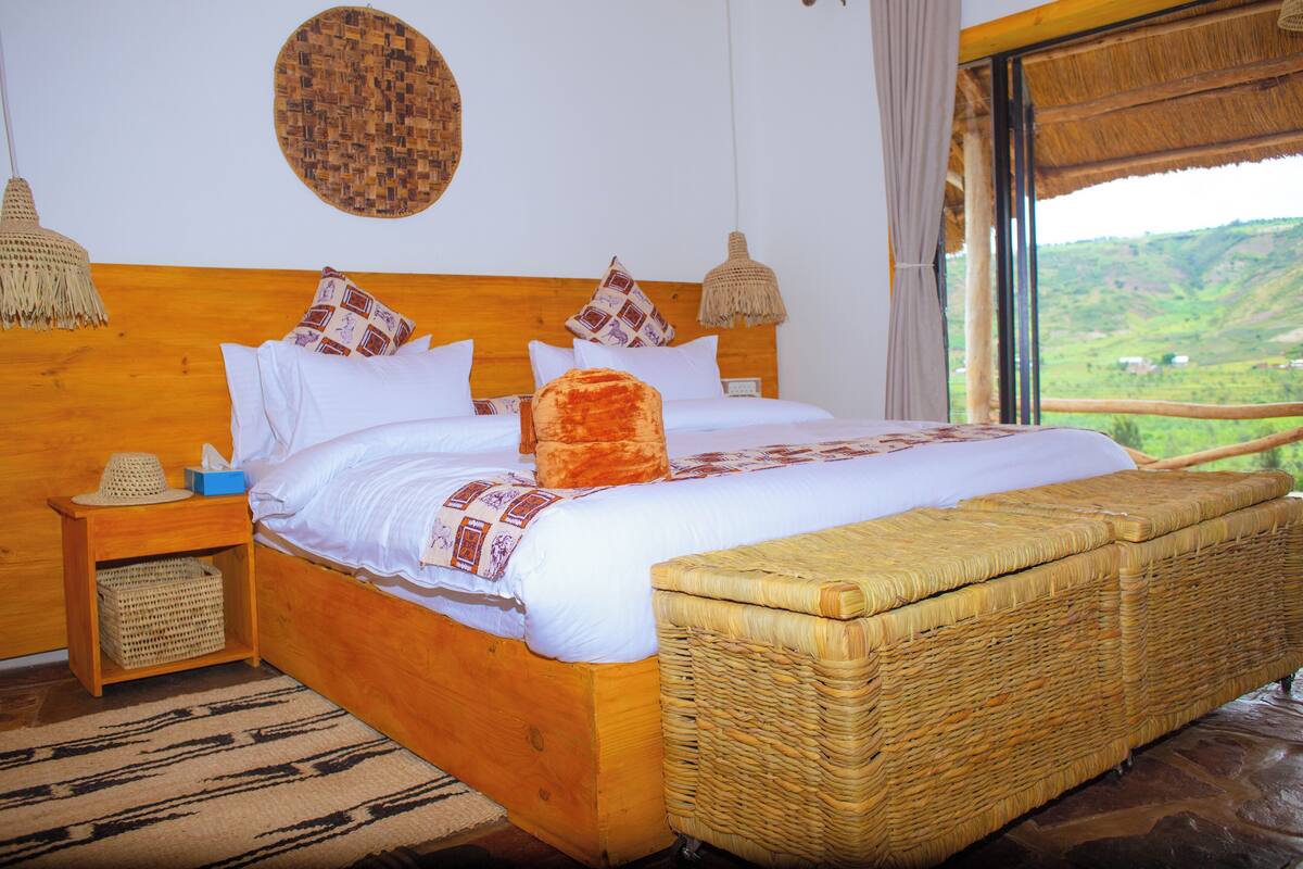 A wooden bed with white bedding, a wicker bench, and a view of a green landscape through a window.