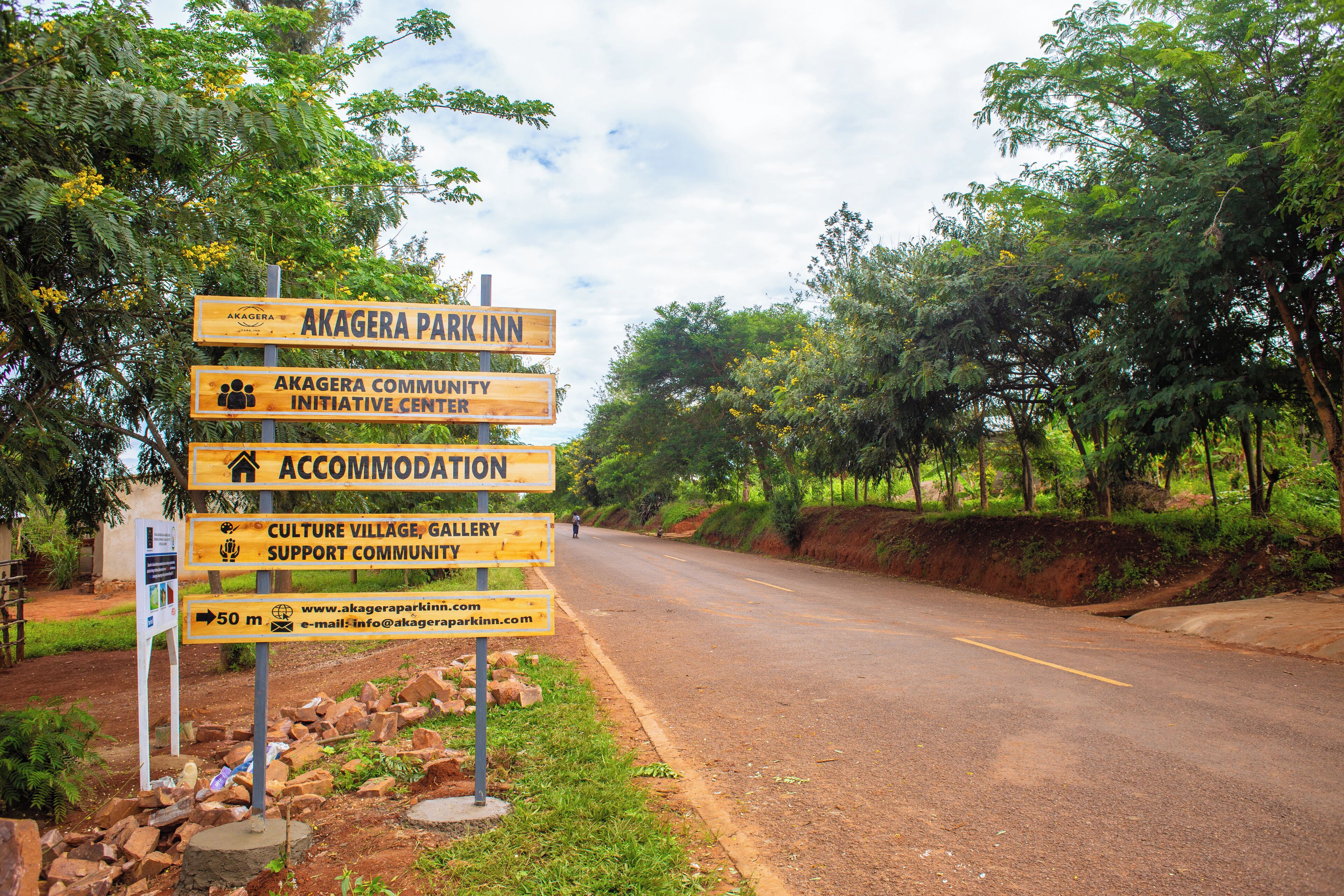 Un panneau indicateur avec de nombreuses directions, notamment vers « Akagera Park Inn », « Akagera Community Initiative Center », « Hébergement » et « Galerie du village culturel ».