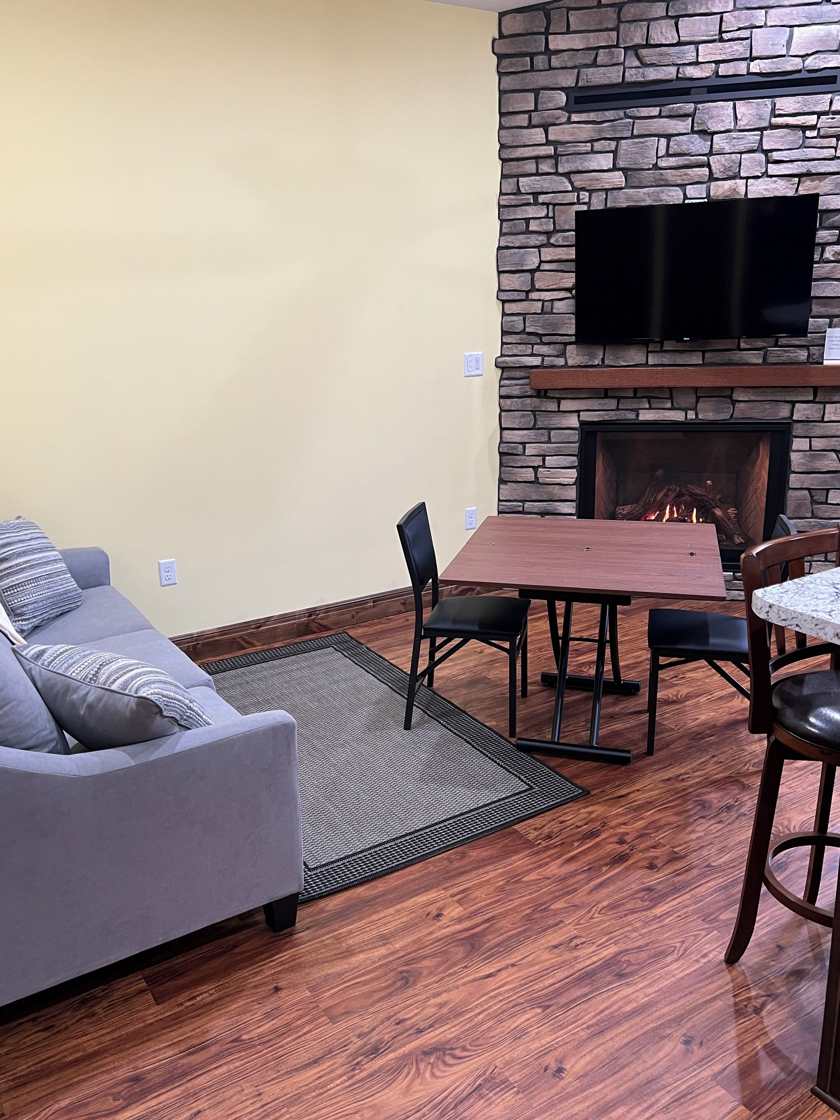 Una sala de estar con chimenea de piedra, un televisor de pantalla plana montado encima, un sofá gris con cojines estampados, una mesa de comedor de madera con sillas y una pequeña mesa redonda con una silla.