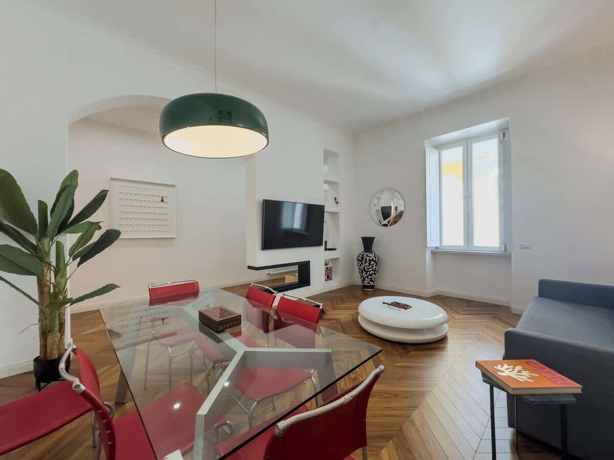 Un salon moderne avec une table à manger en verre, des chaises rouges, un canapé gris, un pouf rond, un téléviseur et une grande suspension verte.