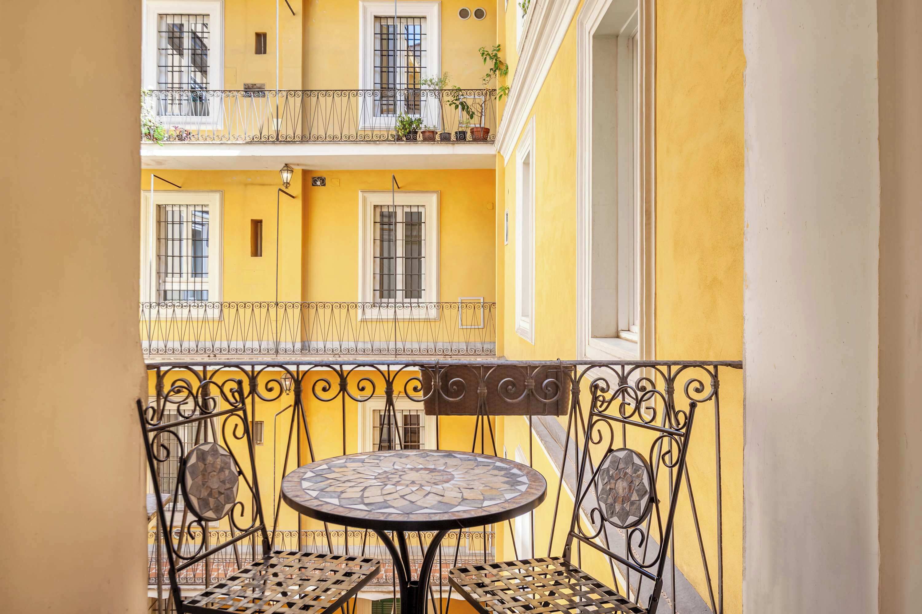 A balcony with a round table and two chairs, a yellow building in the background, and barred windows.