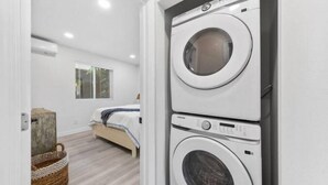 Un lavadero con lavadoras y secadoras apiladas, una cama con colcha a rayas azul y blanca, una cesta de mimbre y una ventana con persianas.