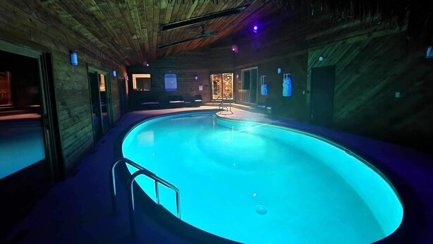 Una amplia piscina interior con techo y paredes de madera, rodeada de zonas de estar y un pequeño bar junto a la piscina.