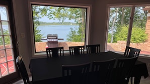 Un comedor con mesa y sillas, ventanas amplias con vista a un lago y una parrilla, además de una pared de ladrillo.