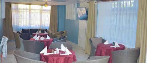 A hotel dining area with round tables covered in red tablecloths, wicker chairs, and a ceiling fan.
