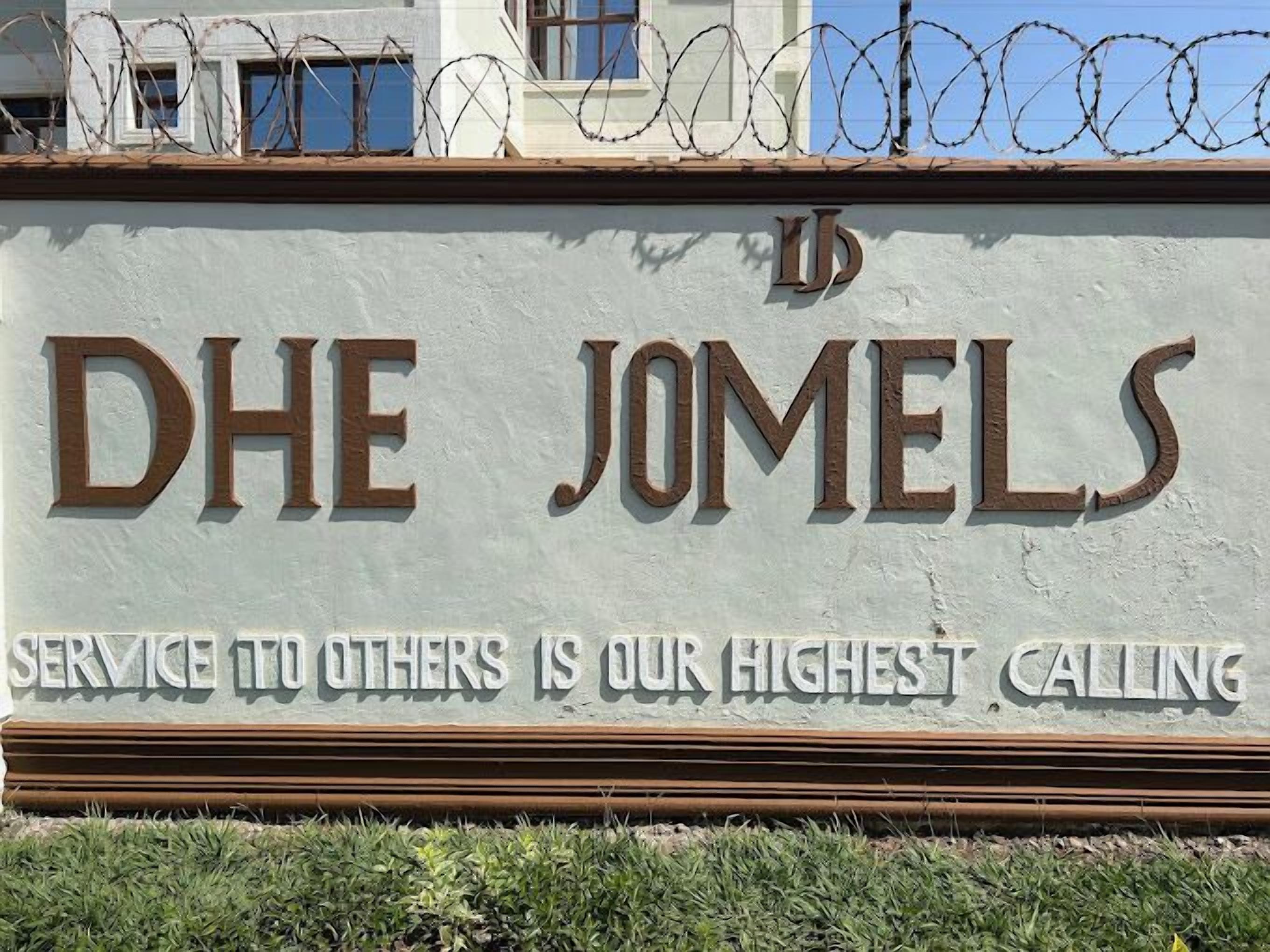 Een bord met de tekst 'DHE JOMELS' en de slogan 'Dienstbaarheid aan anderen is onze hoogste roeping'.