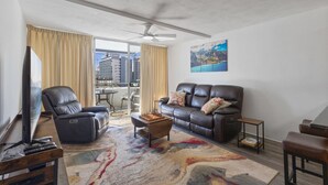 Sala de estar con sofá de cuero, sillón reclinable, mesa de centro y televisor. Balcón con vista a edificios.