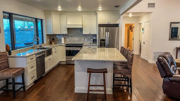 A modern kitchen with white cabinets, a central island with stools, a stainless steel refrigerator, and a window overlooking a garden. - Hubbard Shores (Breckenridge)