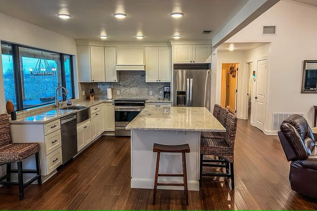 A modern kitchen with white cabinets, a central island with stools, a stainless steel refrigerator, and a window overlooking a garden.