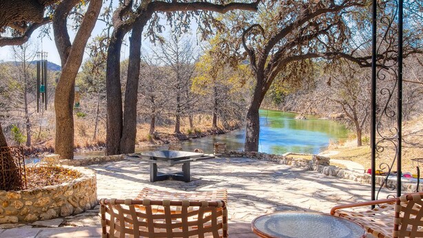 An outdoor patio with a table and chairs, a stone planter, and a view of a body of water. - Luxury Home on 100 acre Secluded Ranch, Waterfront, Little Frio Creek (Leakey)