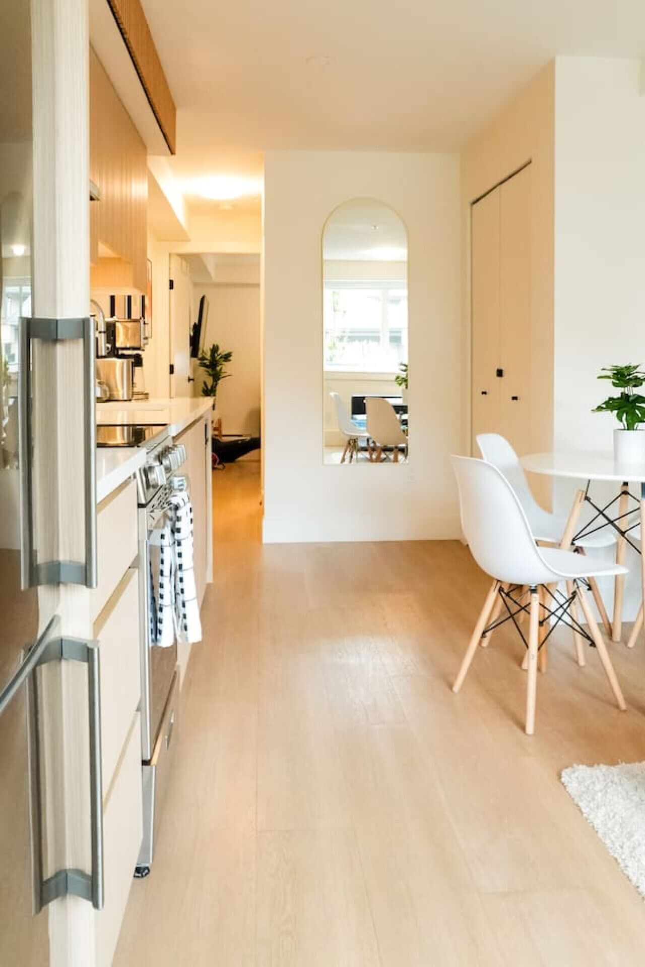 A modern kitchen with stainless steel appliances, a dining area with white chairs and a table, and a hallway leading to a mirrored wall.