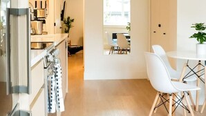 A modern kitchen with stainless steel appliances, a dining area with white chairs and a table, and a hallway leading to a mirrored wall. - Luxury Scandinavian Suite| Cozy and Bright (North Vancouver)