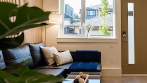 A modern living room with a leather sofa, a glass coffee table with chocolates, and a view of neighboring buildings through a large window. - Luxury Scandinavian Suite| Cozy and Bright (North Vancouver)