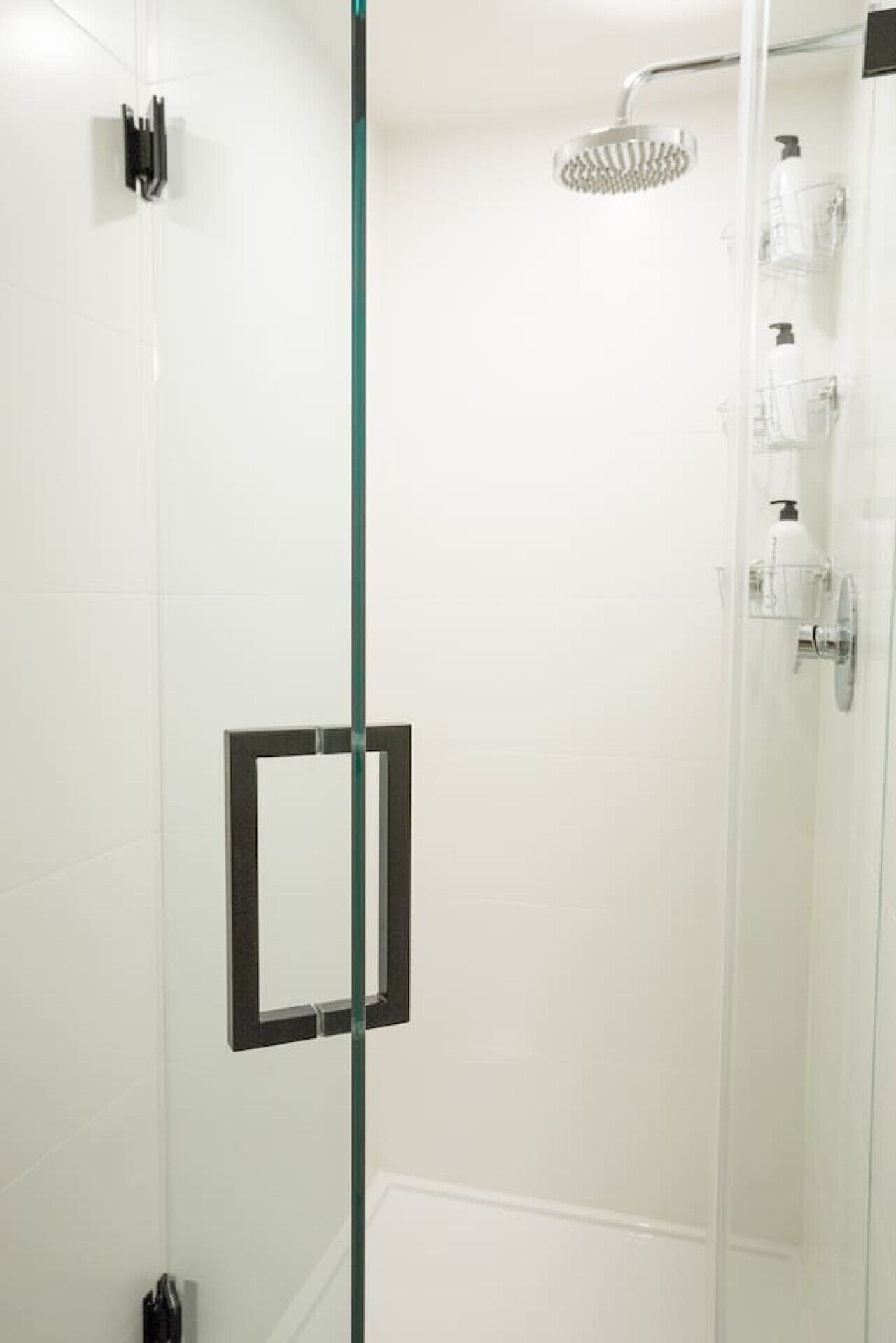 A glass-enclosed shower with a handheld showerhead, a glass shelf with toiletries, and a black-framed mirror.