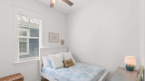 A bedroom with a bed, a bedside table with a lamp, a window with blinds, and a ceiling fan.
