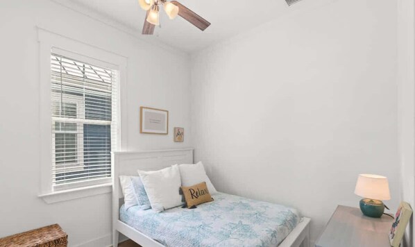 Un dormitorio con una cama, una mesita de noche con lámpara, una ventana con persianas y un ventilador de techo.